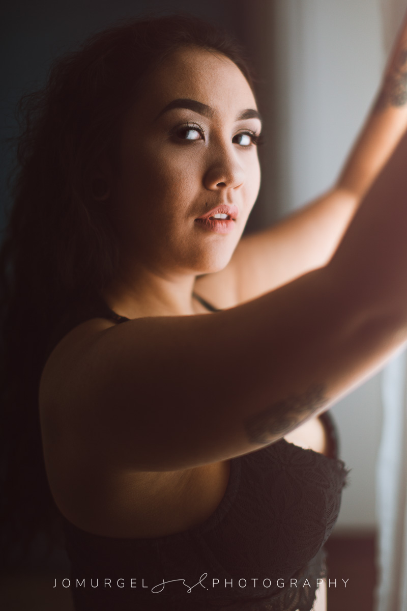 woman in black bra, in front of a window, looking at the camera over shoulder