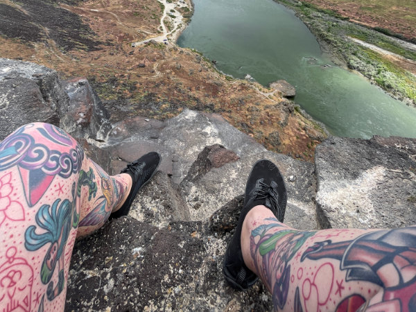 A photo of tattooed legs draped over the edge of a cliff overlooking rocks and the Snake River.